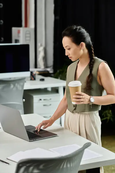 A focused professional reviews work on her laptop while holding a coffee cup in a stylish office. — Stock Photo