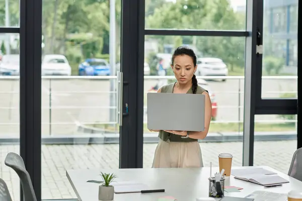 Une femme professionnelle concentrée se tient dans un bureau lumineux, engagée dans son travail avec un ordinateur portable. — Photo de stock