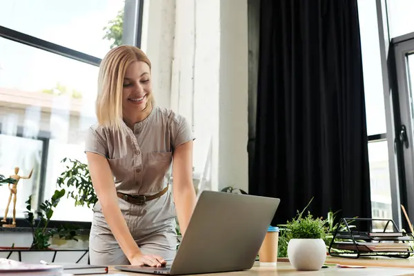 A cheerful young professional engages with her laptop amidst a vibrant office atmosphere. — Stock Photo