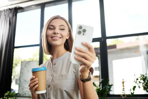 A young professional smiles while holding coffee and checking her phone, immersed in her work. — Stock Photo