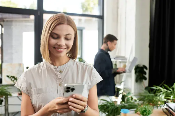 A cheerful coworker smiles while checking her phone, as a colleague focuses on his laptop nearby. — Stock Photo