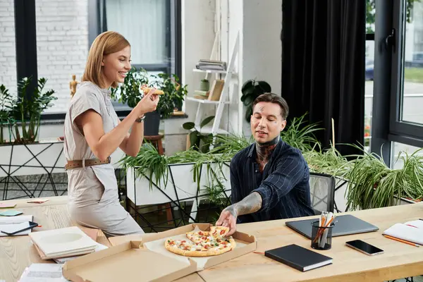 Two young coworkers share a moment as they savor pizza in their vibrant office environment. — Stock Photo