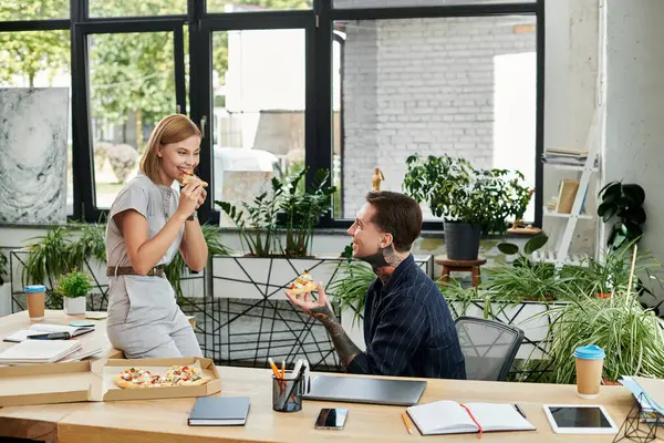 Two coworkers share a cheerful moment over pizza while surrounded by greenery in the office. — Stock Photo