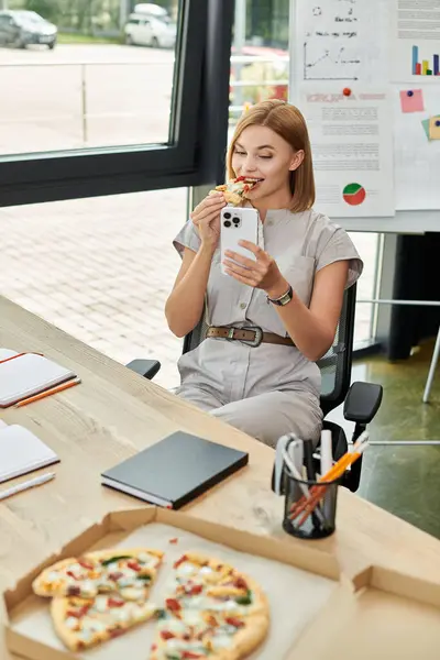 A young woman takes a bite of pizza while checking her smartphone at her office desk. — Stock Photo
