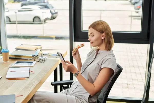 A young professional casually eats pizza and checks her phone in a stylish workspace. — Stock Photo