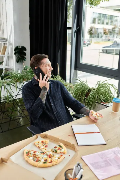 Un joven compañero de trabajo sonríe mientras habla por teléfono, disfrutando de pizza en su escritorio. - foto de stock