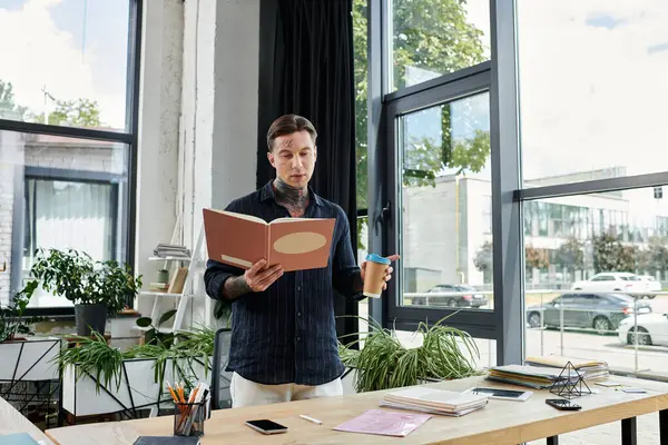 A young coworker stands at a desk, reading from a binder while holding a drink. — Stock Photo
