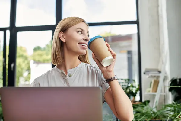 A cheerful young colleague savors coffee while working at her desk in a vibrant office. — Stock Photo