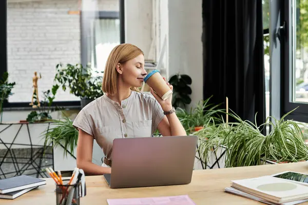 A young professional sips coffee peacefully amidst vibrant plants at her office workspace. — Stock Photo