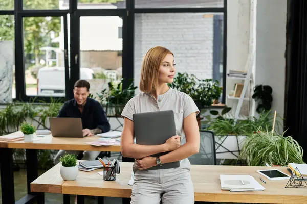 A young professional holds a laptop while her coworker focuses on work at a shared desk. — Stock Photo