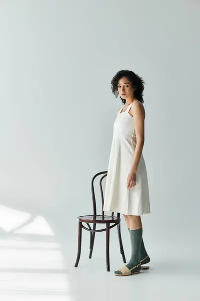 A fashionable woman in chic attire poses gracefully next to a minimalist wooden chair. — Stock Photo