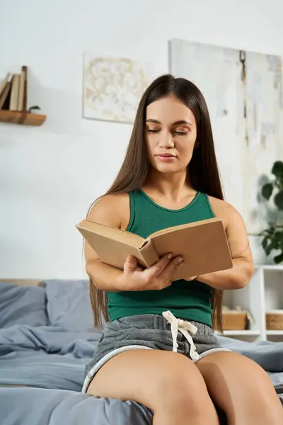 A young brunette woman with short stature relaxes on her bed, fully engrossed in a book during a cozy afternoon at home. — Stock Photo