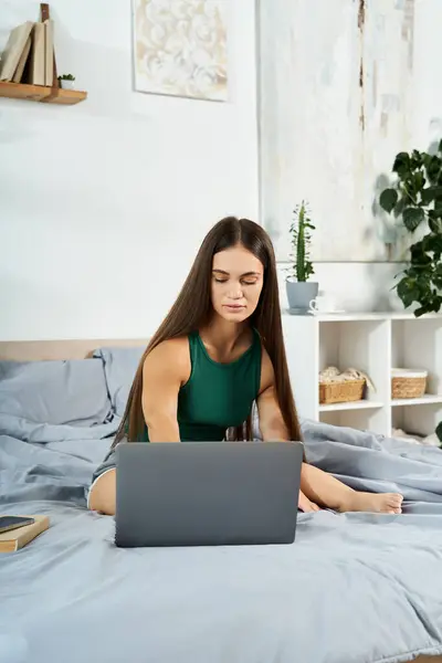 A young brunette woman with short stature is comfortably seated on her bed, engaged with her laptop while enjoying her time at home. — Stock Photo