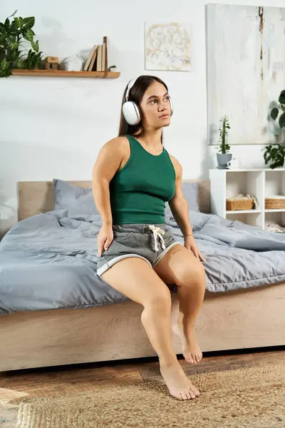 A young brunette woman with a short stature is peacefully relaxing at home, lost in music while sitting comfortably on the edge of her bed. — Stock Photo