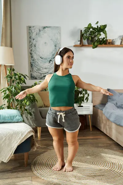 A young brunette woman with short stature dances joyfully in her cozy living room, immersed in music and surrounded by greenery. — Stock Photo