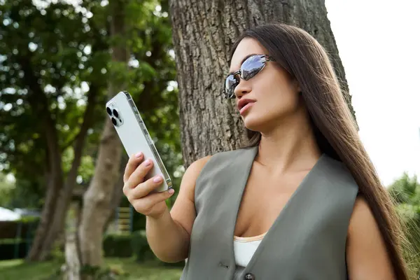 Una giovane donna bruna con una bassa statura vestita con disinvoltura si appoggia ad un albero, assorbita nel suo telefono, godendo l'aria aperta in una giornata di sole. — Foto stock