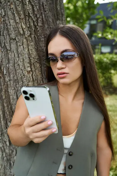 In un ambiente all'aperto vibrante, una donna bruna con una bassa statura si erge accanto a un albero, intensamente concentrata sul suo smartphone in mezzo al verde. — Foto stock