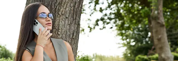 Eine junge brünette Frau mit kurzer Statur steht unter einem Baum und lächelt, während sie an einem sonnigen Tag im Freien telefoniert. — Stockfoto