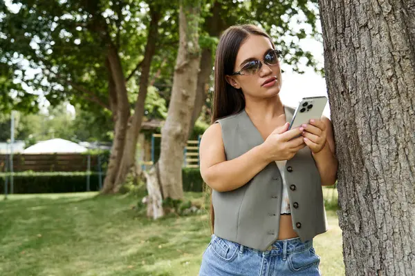 A young brunette woman with short stature stands outdoors, engaged with her smartphone, surrounded by nature's beauty under the sun. — Stock Photo