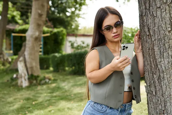 A young brunette woman with short stature stands outdoors, absorbed in texting on her smartphone. She enjoys a tranquil moment surrounded by greenery. — Stock Photo