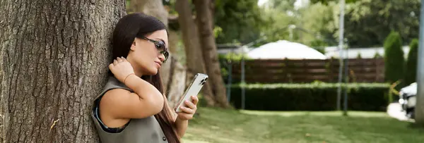A cheerful brunette woman with short stature relaxes outside, leaning against a tree while deeply engaged with her smartphone, soaking up the sunshine. — Stock Photo
