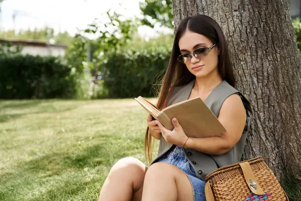 A petite brunette woman with short stature relaxes by a tree, engrossed in her book, enjoying the peaceful atmosphere of the outdoors. — Stock Photo