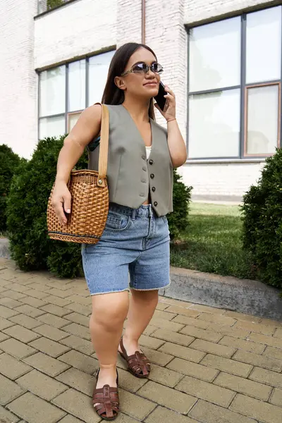 A short-statured brunette woman strolls along a sunny pathway, engaging in a lively phone conversation. — Stock Photo