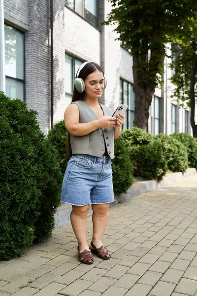 A young brunette woman with headphones strolls leisurely while checking her phone, surrounded by lush greenery. — Stock Photo