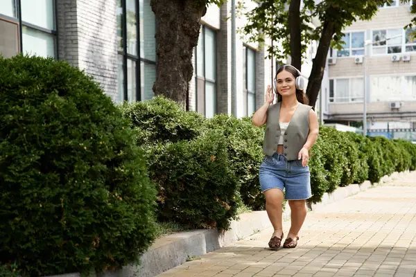 Una joven morena alegre con baja estatura pasea por un sendero bordeado de vibrantes verdor, abrazando el día soleado al aire libre. - foto de stock