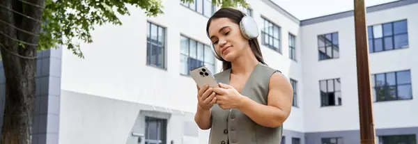A cheerful brunette woman with short stature enjoys her time outdoors, listening to music with headphones while checking her phone. — Stock Photo