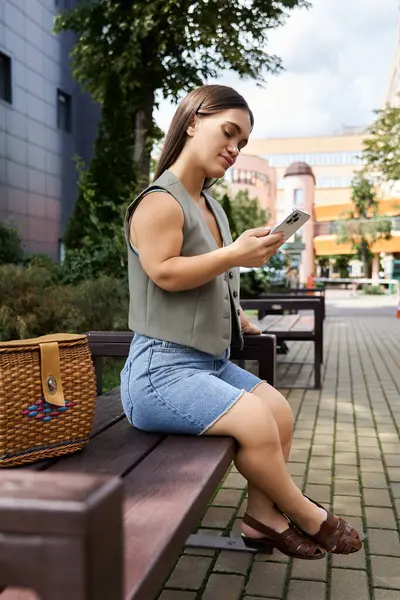 A brunette young woman with a joyful expression sits outdoors, comfortably checking her phone, enjoying the bright day. — Stock Photo