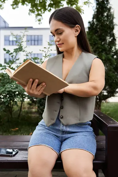 A short-statured brunette woman relaxes on a bench, engrossed in a book under the shade of trees during a sunny day. — Stock Photo