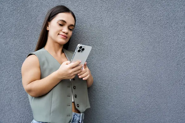 A cheerful brunette woman stands against a wall, engaging with her smartphone on a bright, beautiful day outside. — Stock Photo