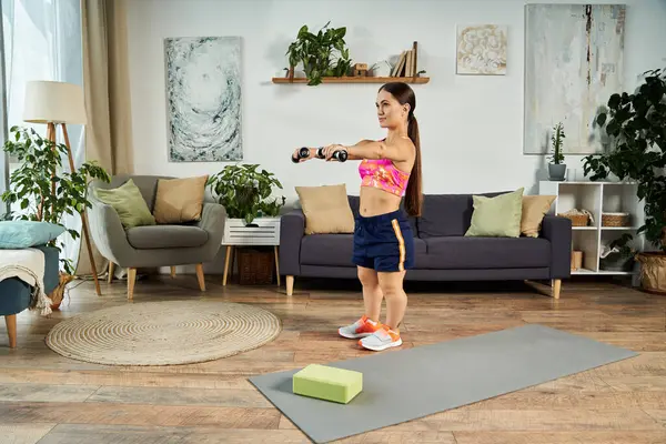 Eine entschlossene brünette Frau mit einer kurzen Statur macht ein Heimtraining, indem sie Gewichte auf ihrer Matte in einem stilvollen Wohnzimmer mit viel Grün benutzt.. — Stockfoto