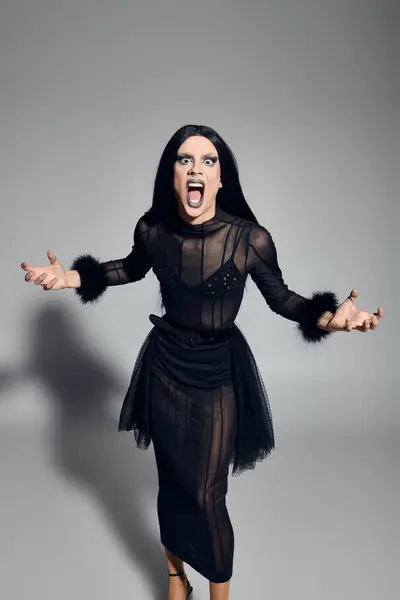 Una llamativa drag queen expresa una energía feroz, completamente adornada en un elegante conjunto negro sobre un fondo gris. - foto de stock