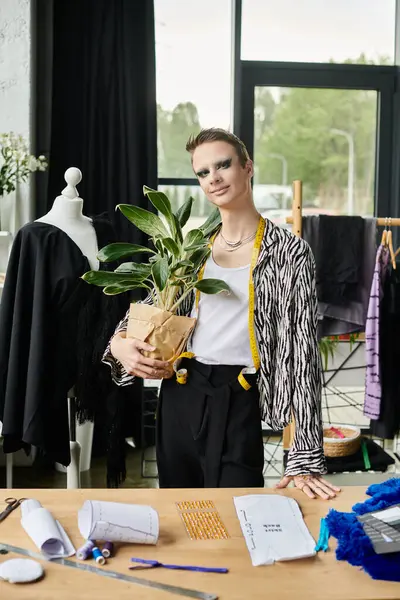 A confident gender fluid designer holds a potted plant while surrounded by artistic materials and garments. — Stock Photo
