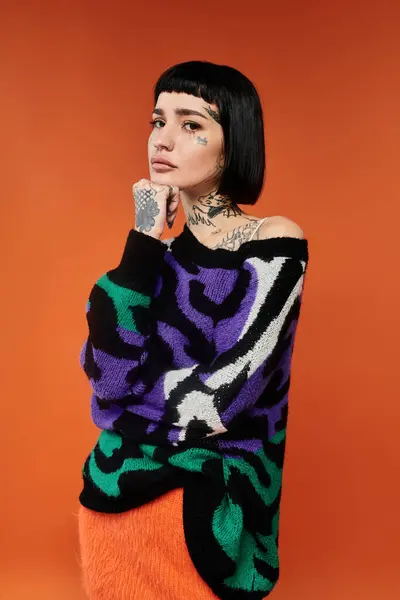 Una joven posa con confianza, mostrando sus tatuajes y suéter vibrante sobre un llamativo fondo naranja. - foto de stock