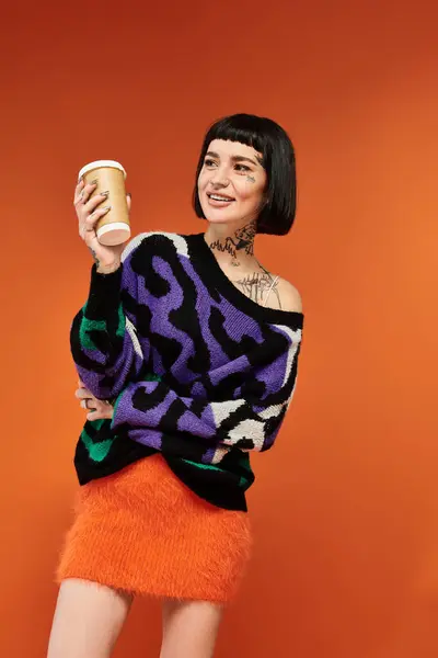 A young woman with short hair and colorful tattoos smiles brightly while holding a coffee cup in a studio setting. — Stock Photo