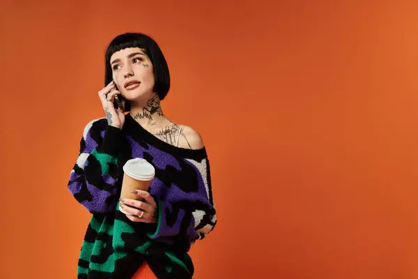 A young woman with short hair and tattoos poses confidently in a colorful sweater while holding a coffee. — Stock Photo