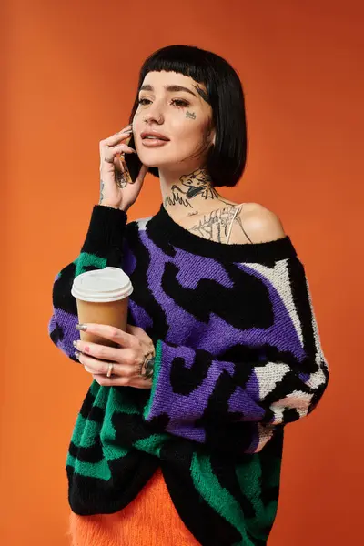 Una mujer joven y elegante con el pelo corto y tatuajes sostiene una taza de café mientras posa contra una pared naranja vívida. — Stock Photo