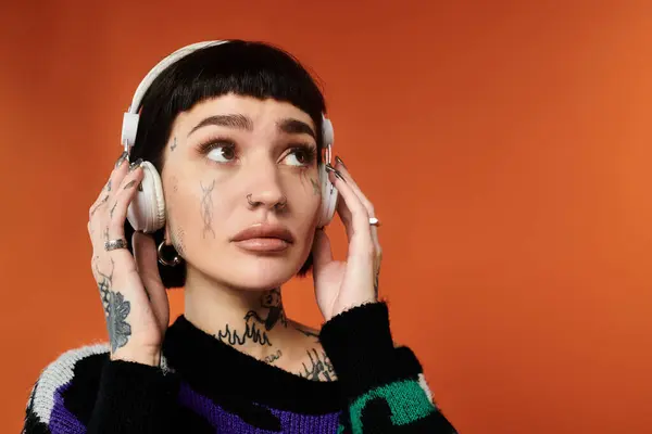 Une jeune femme dans un pull coloré et tatouages écoute de la musique, perdue dans l'instant sur fond orange. — Photo de stock