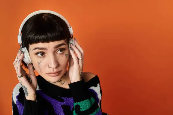A young woman with short hair and tattoos captivates the studio with her bright sweater while listening to music. — Stock Photo