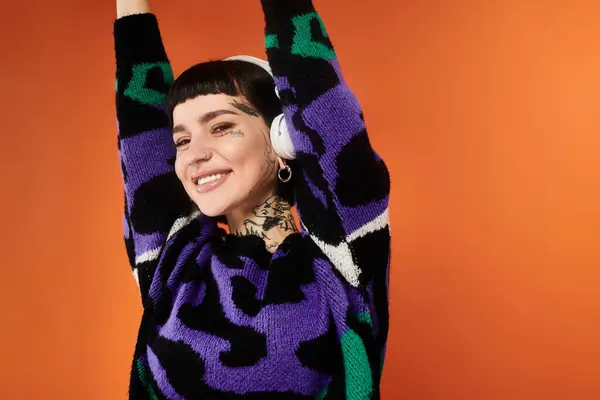 Una giovane donna con un'acconciatura alla moda posa gioiosamente con le braccia alzate, in mostra i suoi tatuaggi e maglione colorato. — Foto stock
