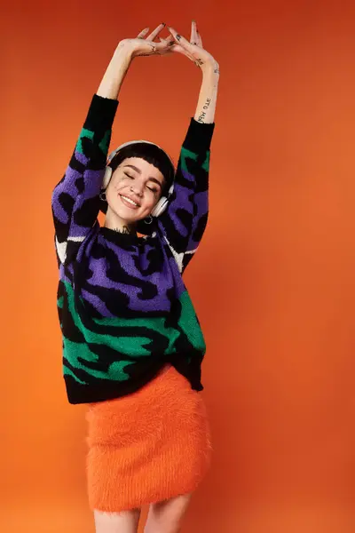 A tattooed woman with short hair smiles, stretching her arms in a colorful sweater against an orange backdrop. — Stock Photo