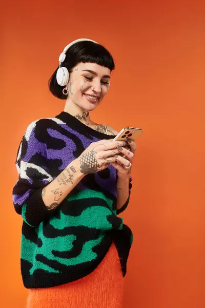 Eine kurzhaarige, tätowierte Frau lächelt auf ihrem Handy in einem hellen Pullover vor orangefarbenem Hintergrund. — Stockfoto