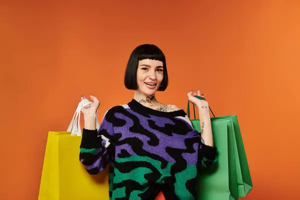 A young woman confidently poses with shopping bags, showcasing her vibrant sweater against a bold orange backdrop. — Stock Photo
