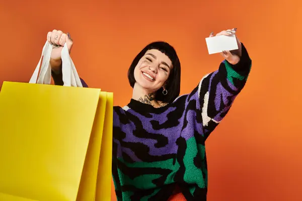 A young woman with short hair and bright tattoos smiles, posing with shopping bags against a colorful backdrop. — Stock Photo