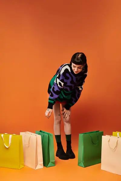 A young woman with short hair and tattoos crouches beside bright shopping bags in a lively studio. — Stock Photo