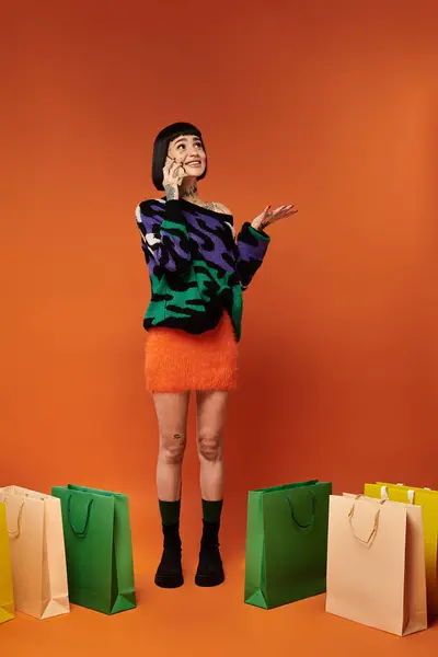 A young woman with short hair and tattoos playfully gestures while surrounded by colorful shopping bags. — Stock Photo