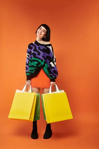 Une jeune femme présente son pull vibrant et ses tatouages uniques tout en tenant joyeusement des sacs à provisions. — Photo de stock